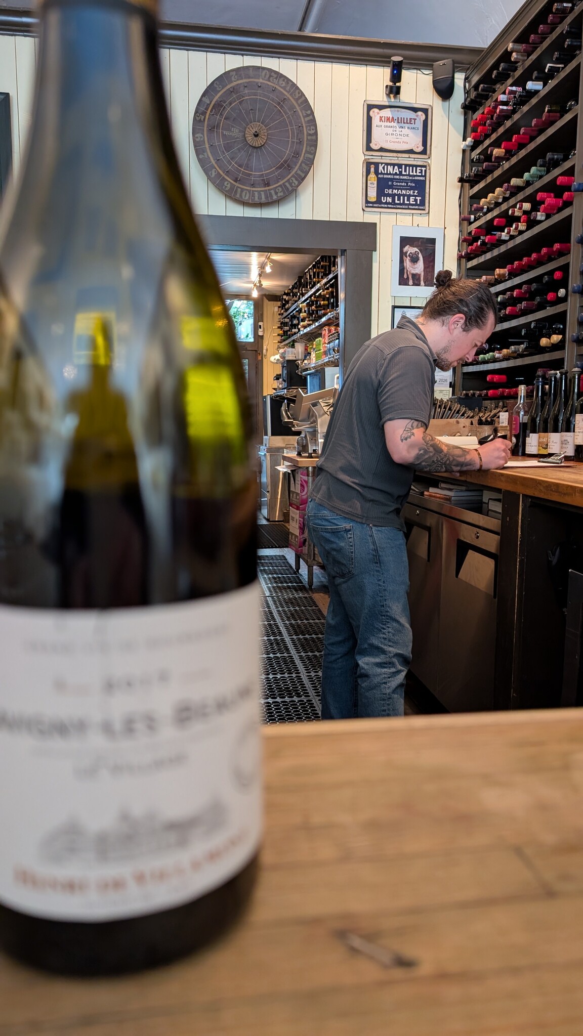 Picture of a wine bar, wine bottle is on the counter and out of focus and the man bent over the counter is writing something. There's many wine bottles on the shelves