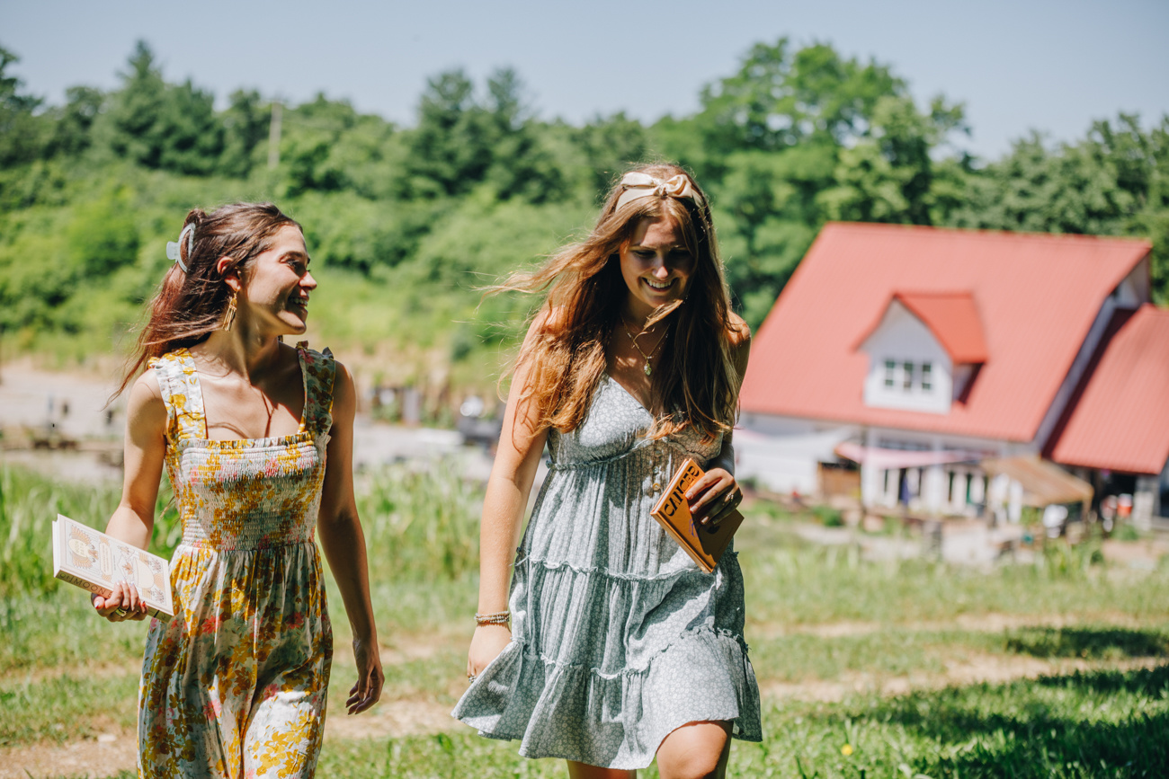 Close up of two women walking with books in hand, one wearing a blue floral dress and the other wearing a yellow full length dress. In the background it a red farm house and lush greenery
