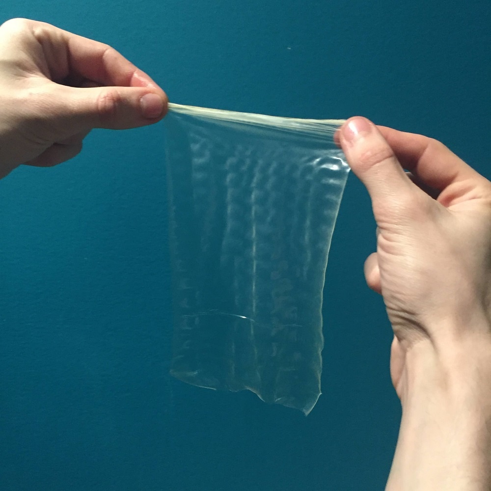 The hands hold up the unrolled condom, now a square sheet of latex.