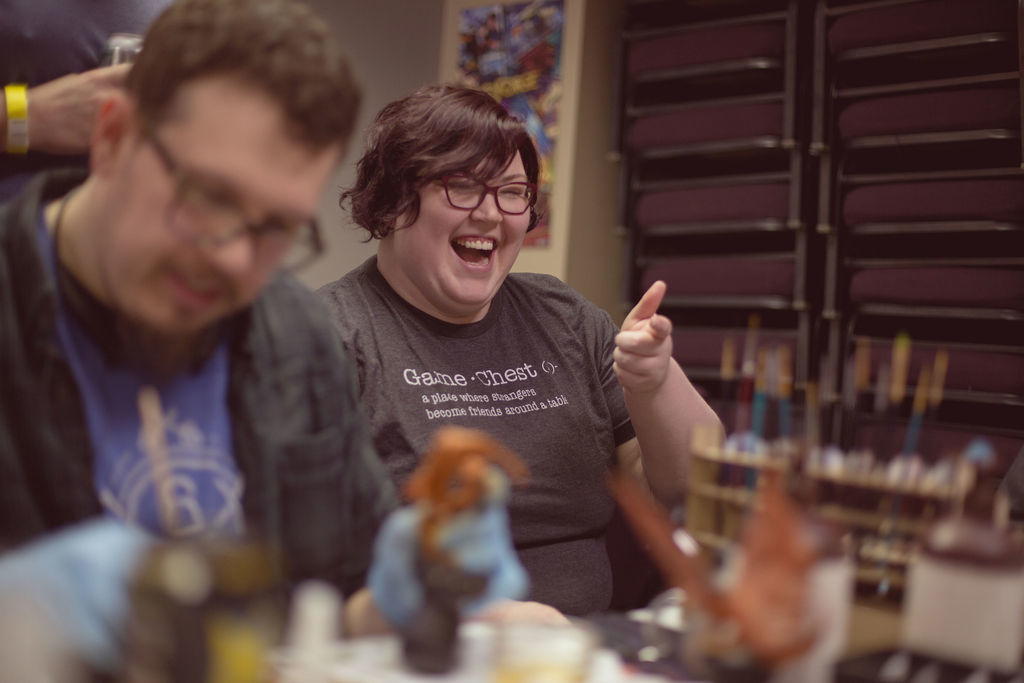 Photo of Amanda in focus shooting finger guns, and Ben out of focus in foreground painting miniatures.