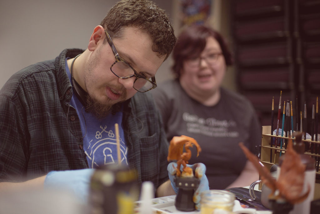 Photo of Ben in foreground painting a wyrm miniature. Amanda blurred in background making conversation.