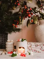 A lit white candle warmer with wax melts, matches, and a match holder sits on marble next to a floral arrangement, a decorative box, and the Mimi & August Mini Aroma Diffuser. A hand lights a tealight under the Mimi & August Mini Aroma Diffuser, s