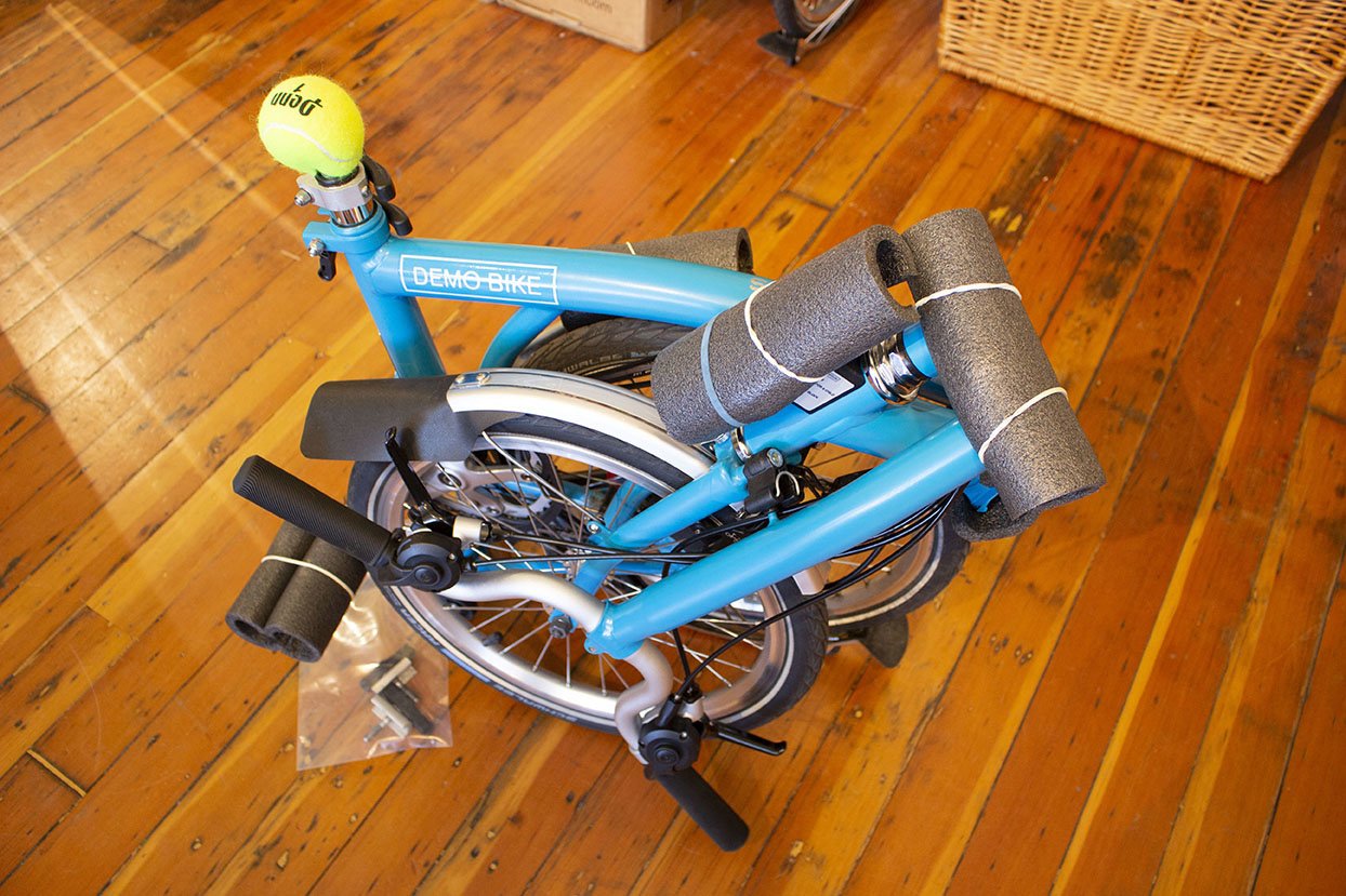 A folded Brompton bicycle with pieces of gray foam pipe insulation covering the pedals and hinges and a tennis ball where the saddle would be