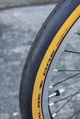 Black tire with light tread, tan sidewall, and Schwalbe One logo on a silver rim with black spokes