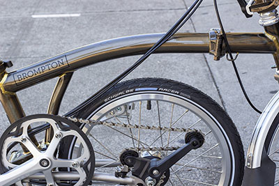 A black tire with a Schwalbe Marathon Racer logo on it on a raw lacquer Brompton in kickstand mode