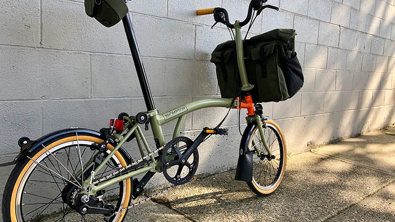 A light green Brompton folding bike with an orange accent and large bag on the front