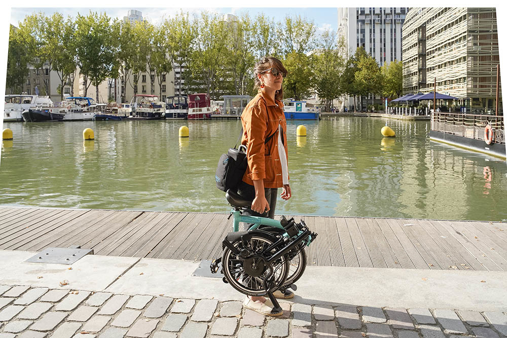 A person with long hair pulled back and an orange jacket, standing on a cobblestone street next to water with boats in it, holding a folded up bicycle by the saddle with one hand