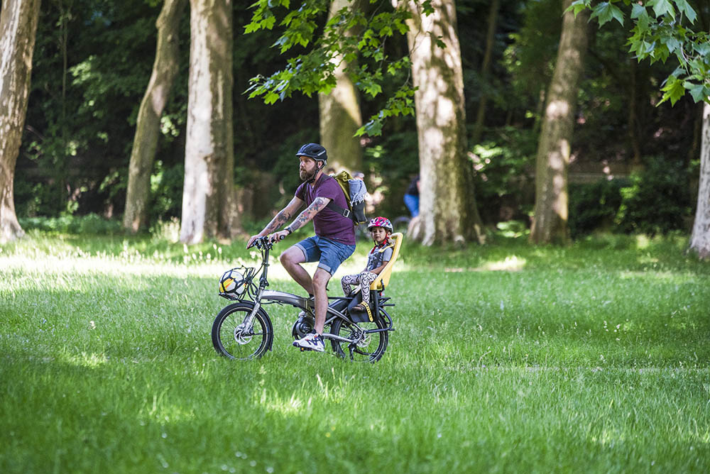 A person with tattooed arms wearing a purple shirt, denim shorts, and a black helmet riding a bike while carrying a small passenger in a kids' seat 