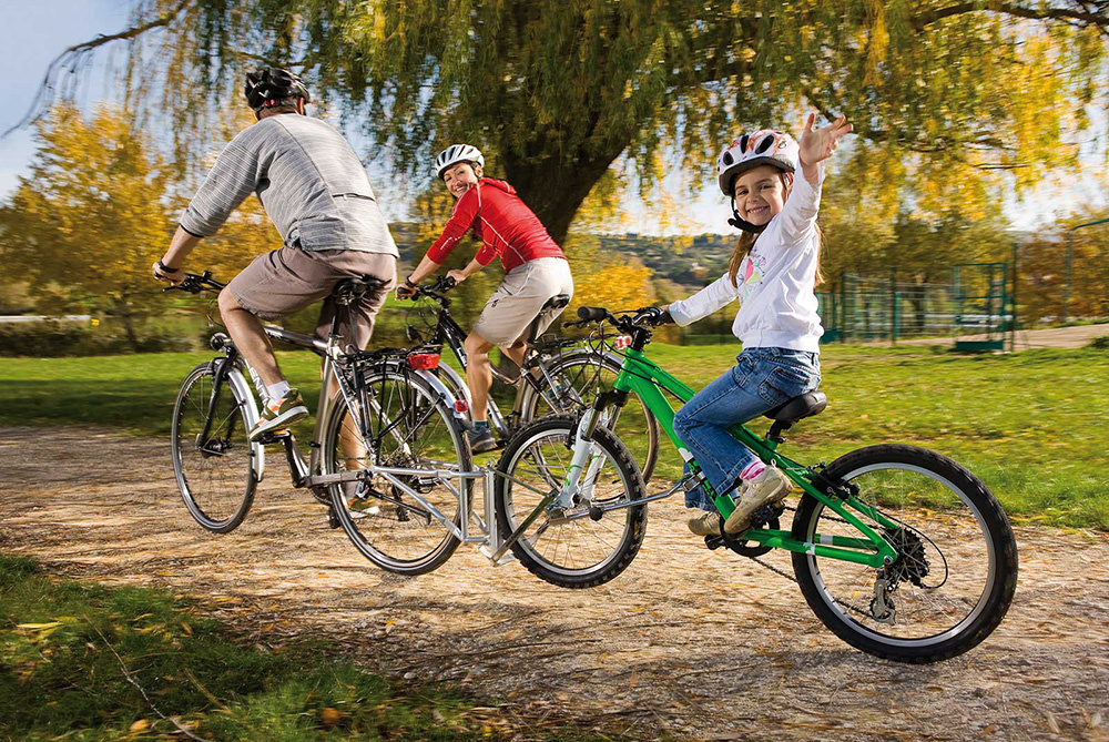 An adult on a bicycle towing a child on a bicycle, the two bikes connected by a metal coupling