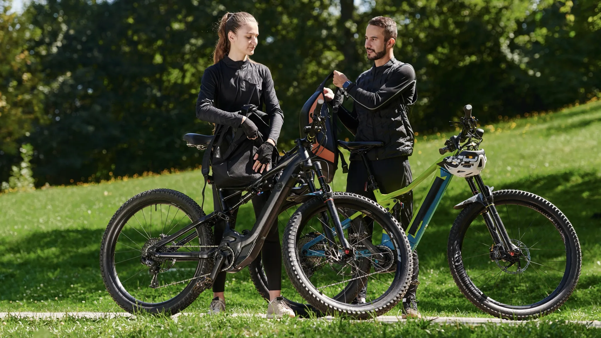 Deux cyclistes préparent leur sac à dos à côté de vélos de montagne électriques dans un parc ensoleillé.