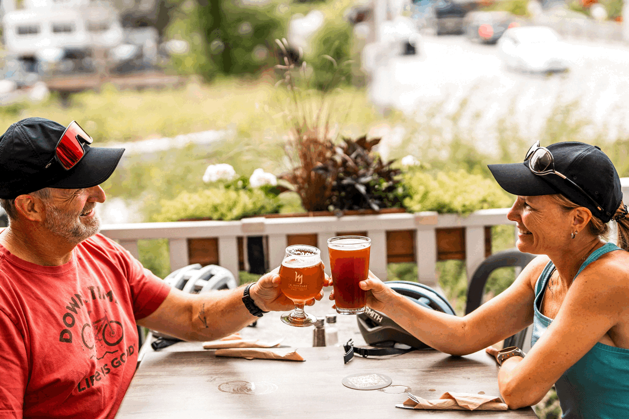 Couple de cyclistes trinquant avec des bières artisanales sur une terrasse ensoleillée après une balade à vélo au Québec.