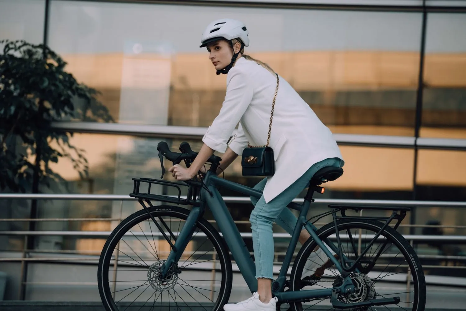 Jeune femme élégante en veston blanc et casque de vélo, roulant sur un vélo électrique urbain devant une façade vitrée moderne.