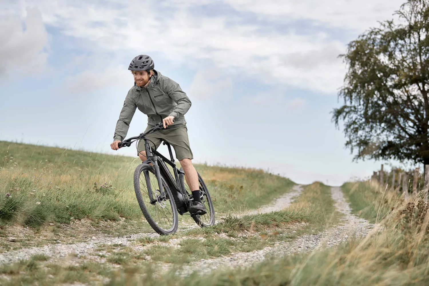 Homme souriant en vélo de montagne électrique, roulant sur un sentier de gravier en pleine nature sous un ciel partiellement nuageux.