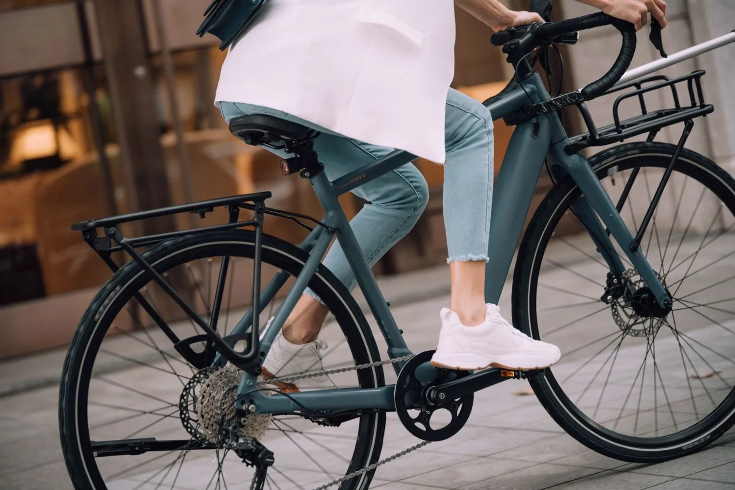 Gros plan sur un vélo électrique urbain de couleur anthracite, pédalé par une femme vêtue d’un manteau blanc et de jeans pâles, en milieu citadin.