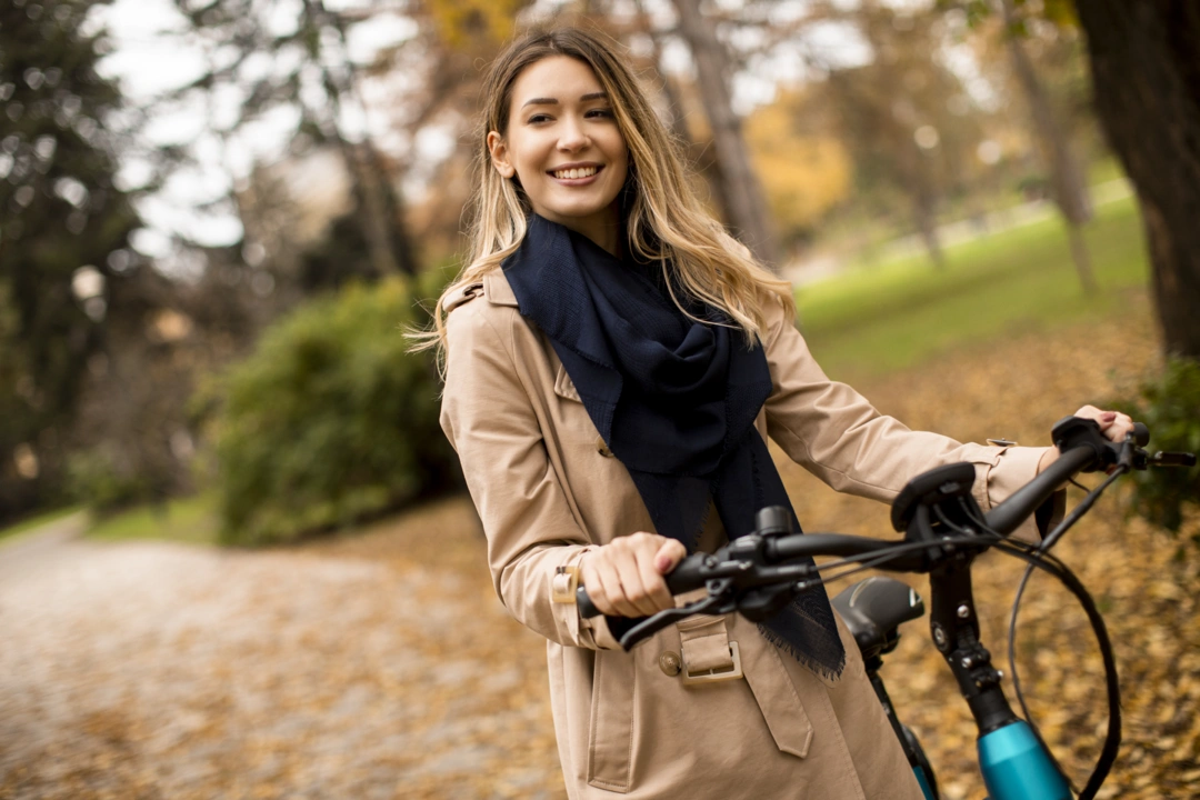 Jeune femme souriante en manteau beige et foulard bleu marine posant avec son vélo électrique dans un parc en automne.