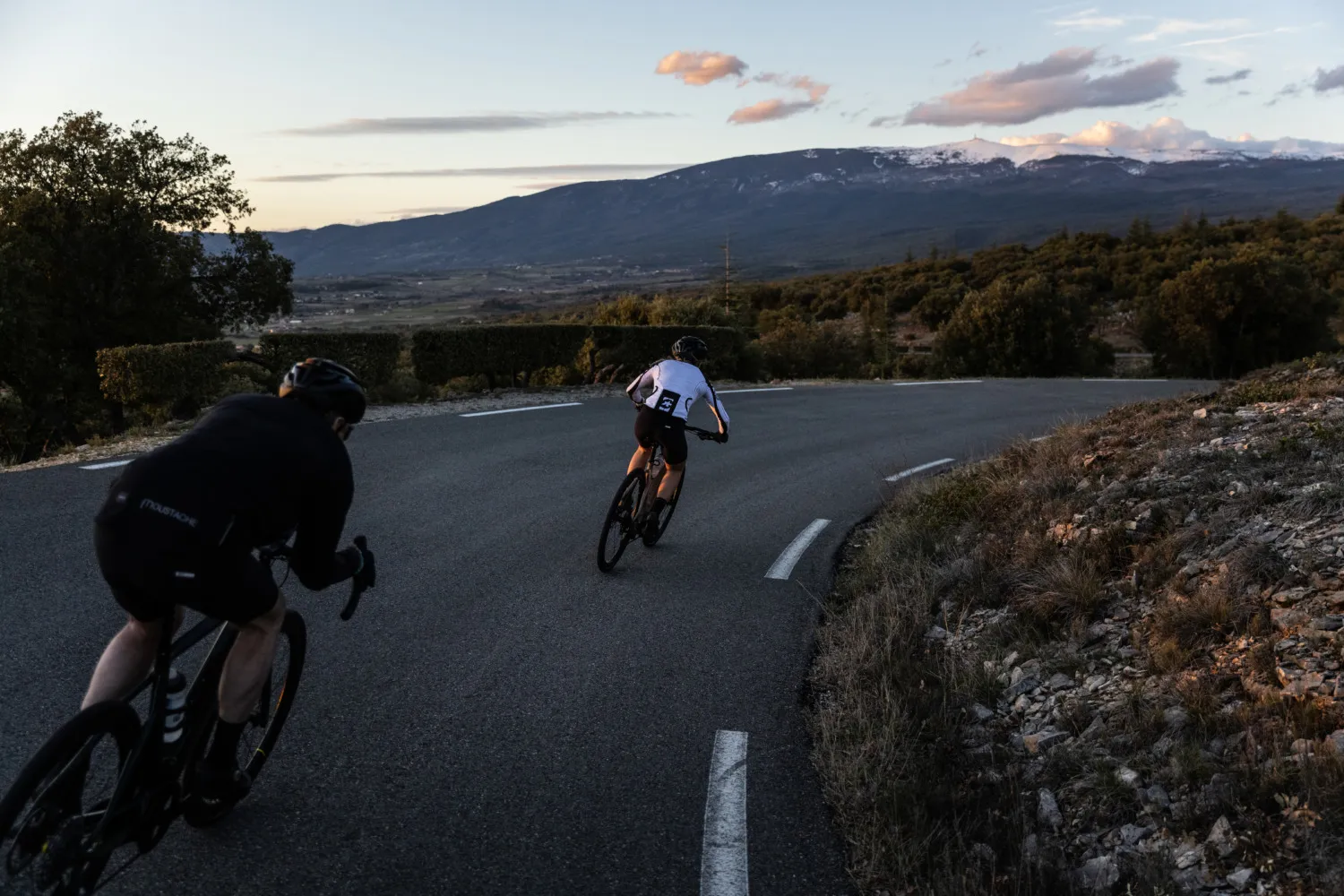 Deux cyclistes en pleine descente sur une route de montagne, au coucher du soleil, avec vue dégagée sur des montagnes.