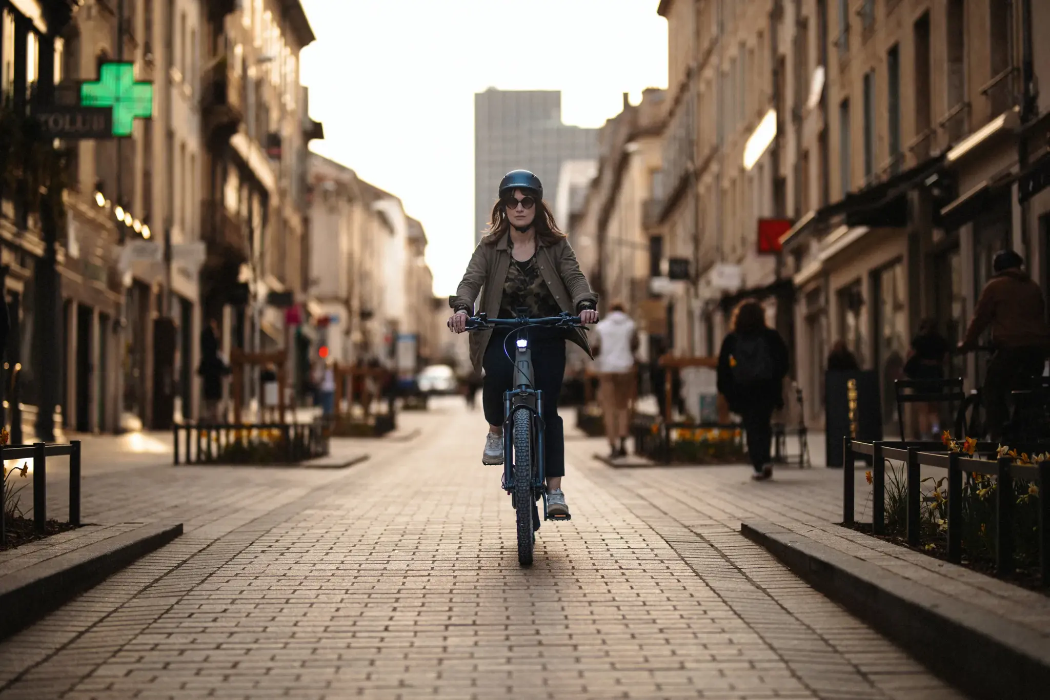Cycliste urbaine portant un casque noir roulant en vélo électrique sur une rue pavée en fin de journée.