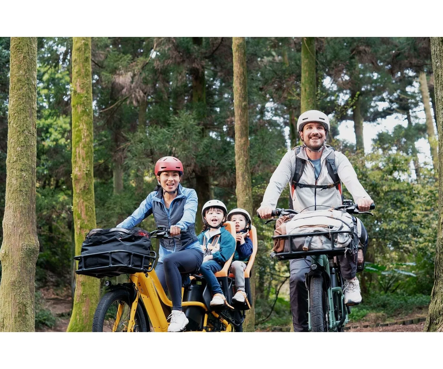 Famille souriante roulant en forêt sur deux vélos électriques cargo, avec deux enfants installés à l’arrière en pleine nature.