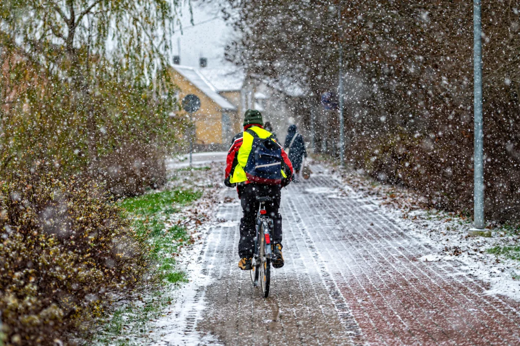 Cycliste en manteau réfléchissant roulant sur une piste cyclable enneigée en hiver, sous une chute de neige abondante.
