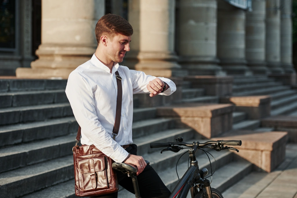 Homme équiper en termes de vêtements et accessoires consultant sa montre devant un bâtiment en pierre, à côté de son vélo de ville stationné.