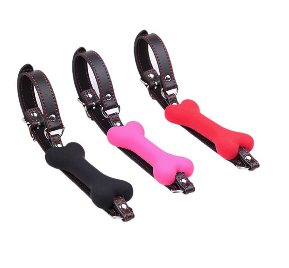 Three bone gags against a white background. The gags are made with silicone and leather straps. One bone is black, another bone is pink, and the other is black. The mouth gags have a metal buckle on the back to adjust the size.