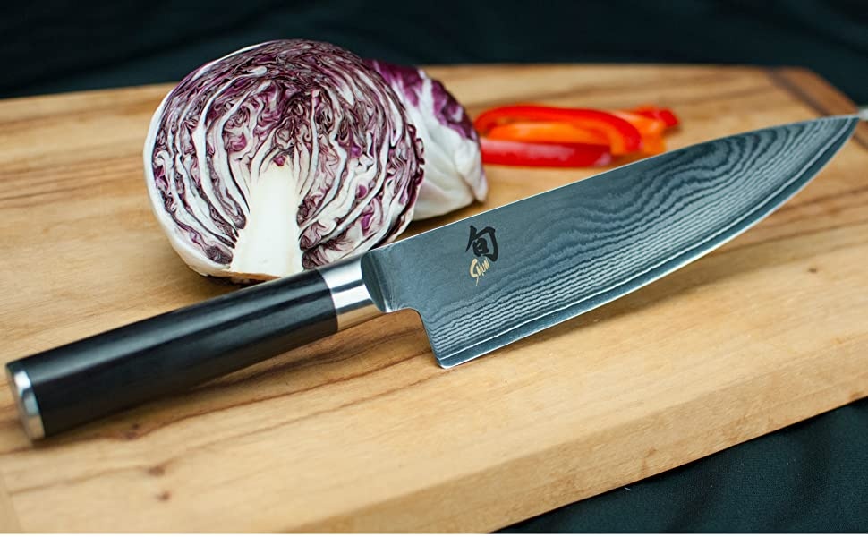 A Shun chef's knife sits on a wooden cutting board next to vegetables. 