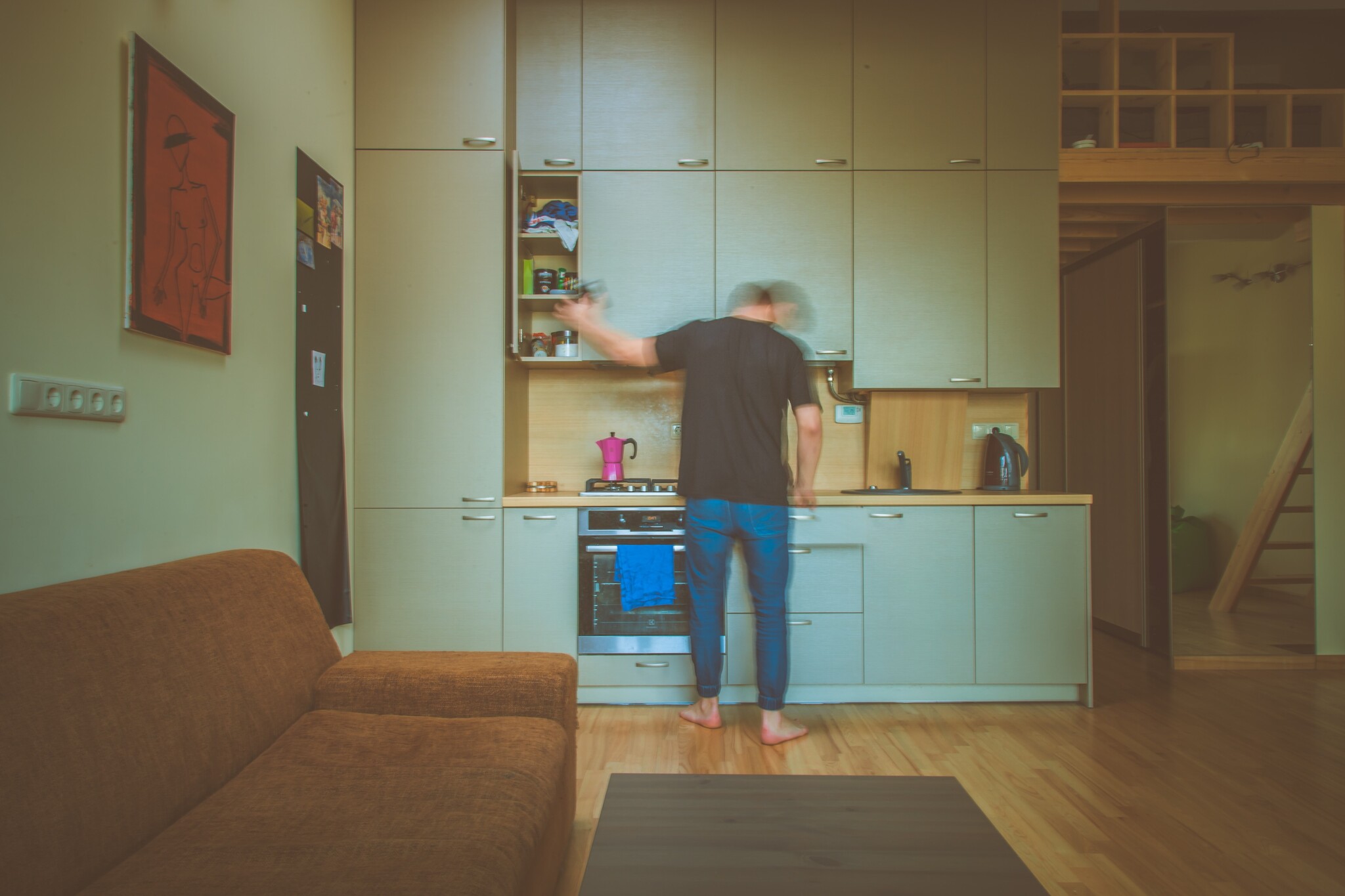 A man stands with his back turned in front of a small kitchen with green cabinets. 