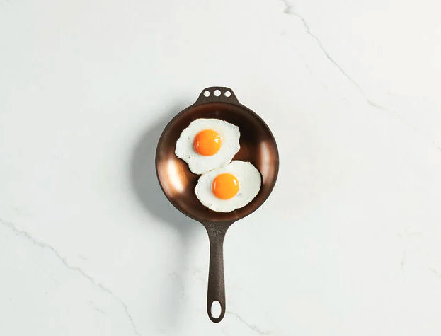 A stainless steel frying pan sits on a white countertop with two fried eggs inside of it. 