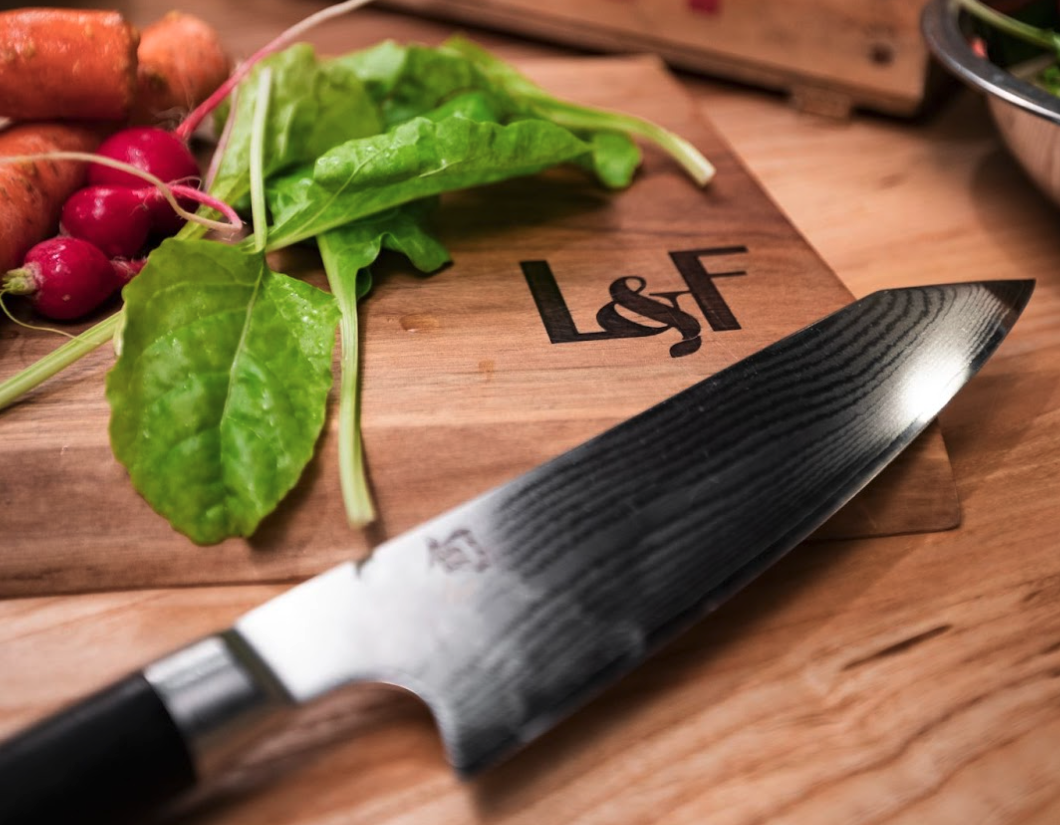 A Shun knife sits on a wooden cutting board with other colorful vegetables. 