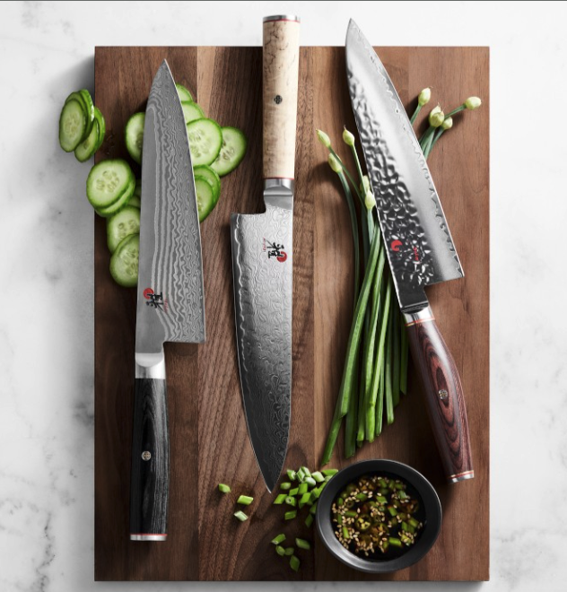 A set of Miyabi chef's knives on a cutting board with sliced cucumbers, green onions, and a soy sauce blend. 