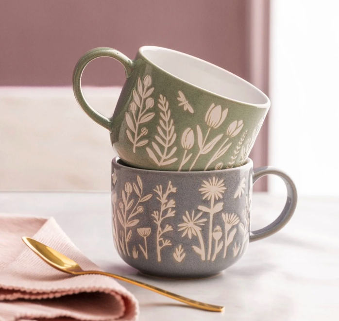 Mason Cash green and blue mugs sit stacked on top of each other on a white surface with a pink towel and golden teaspoon sitting beside them. 