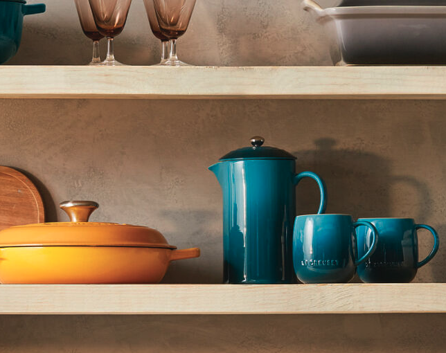 A blue Le Creuset French press sits on a wooden shelf with two blue coffee mugs to its right and an orange braiser to its left. 