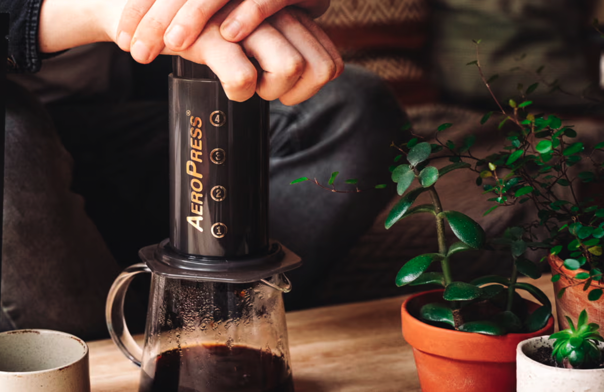 A black aeropress brewer brews coffee into a clear coffee mug on a wooden table with leafy green potted plants beside it. 