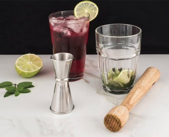 A silver jigger, wooden muddler, a small glass filled with limes and mint, a large glass filled with red liquid, half a lime, and a sprig of mint sit on a white countertop. 