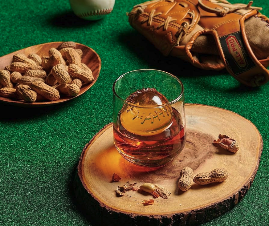 A baseball, baseball glove, bowl of peanuts, and a cup containing a baseball ice sphere and amber liquid sit on astroturf. 