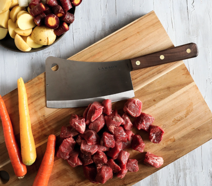 Lamson's Heavy-Duty Meat Cleaver sits on a wooden cutting board with diced red meat and whole orange-and-yellow carrots beside it. In the top left corner, a bowl filled with diced red carrots and potatoes sits on a white countertop. 