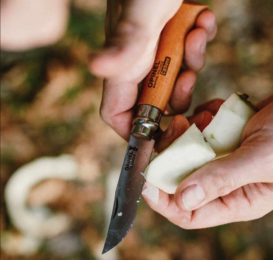 Hands hold a folding Opinel carbon steel knife, using it to slice into a piece of white fruit. 