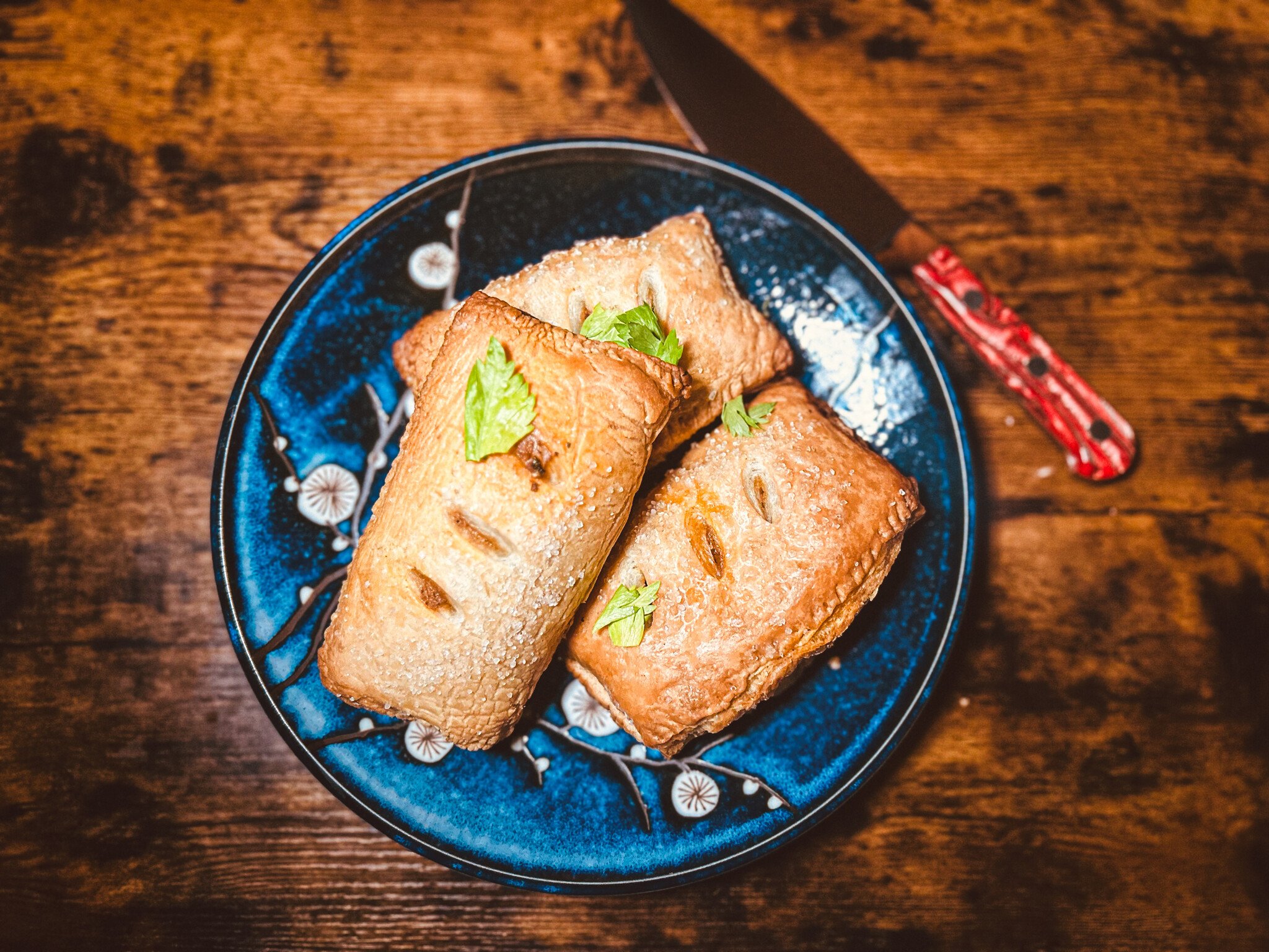 Three puff pastry meat pies sit on a blue plate, which rests on a wooden surface with a red-handled knife beside it. 