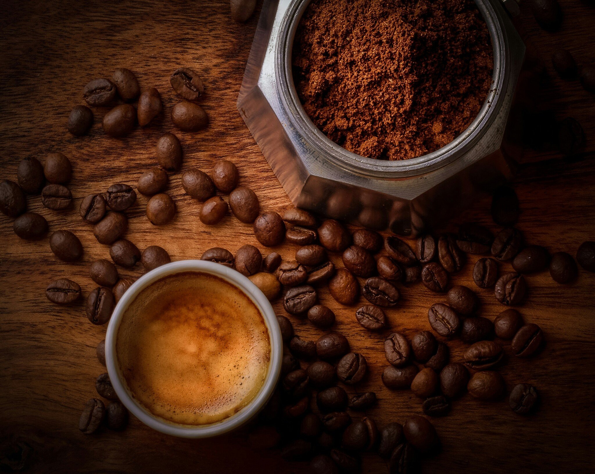 A moka pot filled with ground coffee sits next to a shot of espresso. Both sit on a brown wooden tabletop covered in coffee beans. 