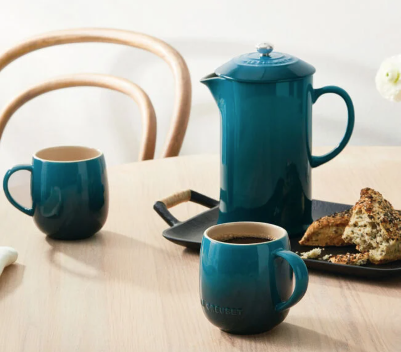 A teal Le Creuset French press sits beside two teal coffee mugs and a tray with a pastry on a wooden table. 