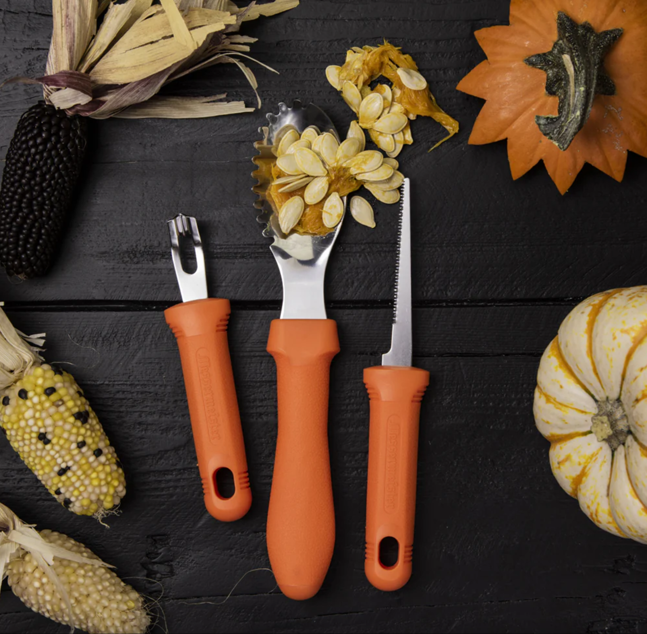 A scraper, a small saw blade, and a scooper with pumpkin guts all sit side by side on a black tabletop surrounded by pumpkins, corn, and fall foliage.