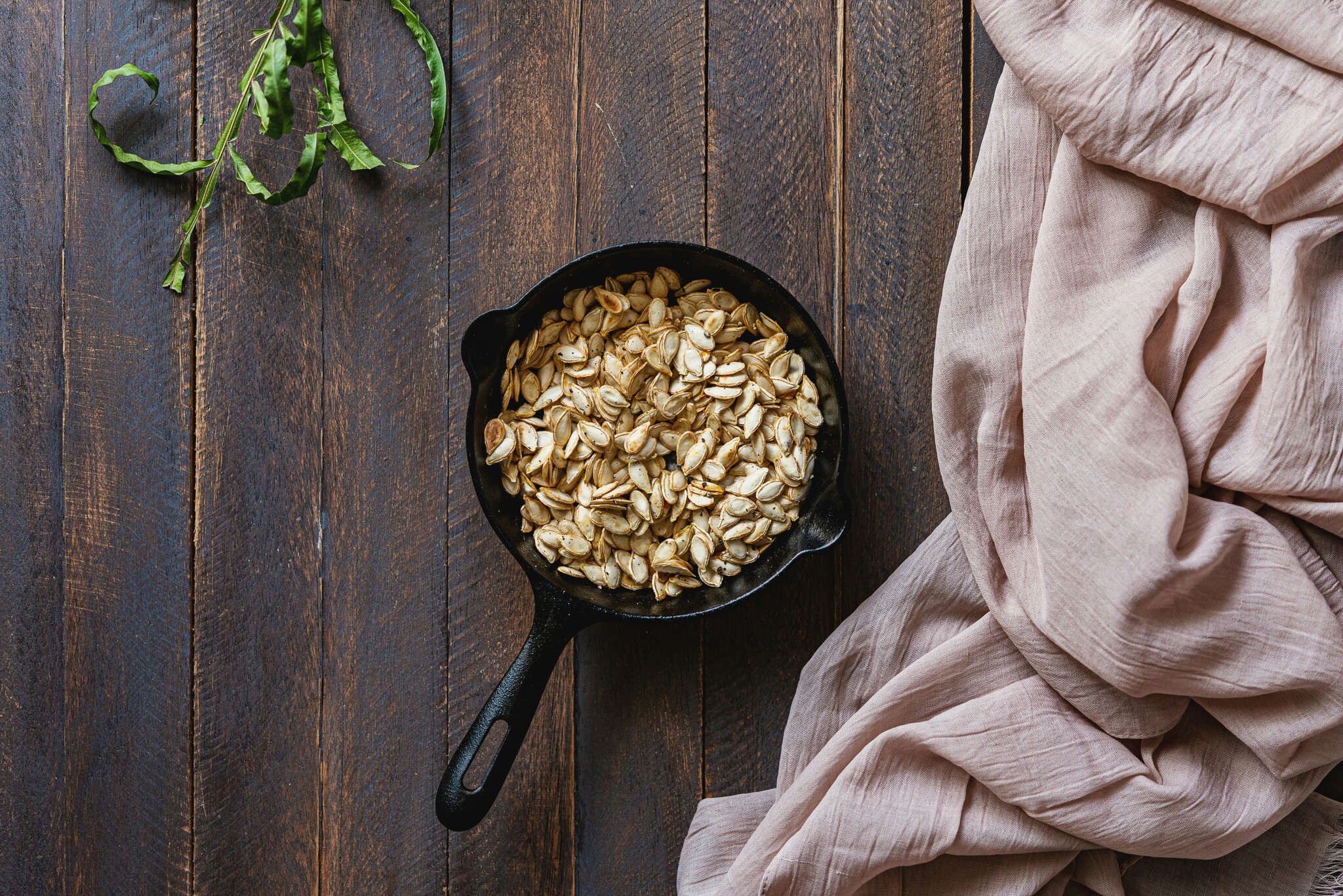 roasted pumpkin seeds sit in a cast iron skillet on a wooden tabletop next to a vine of green herbs and a tan cloth. 