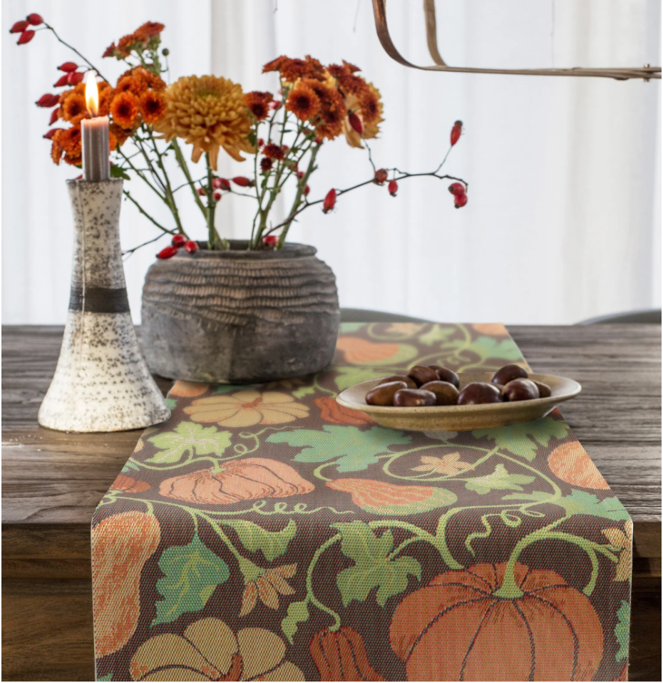 A pumpkin-themed table runner sits on a wooden table with a fall candle, autumnal flowers, and a plate of nuts on top of it. 