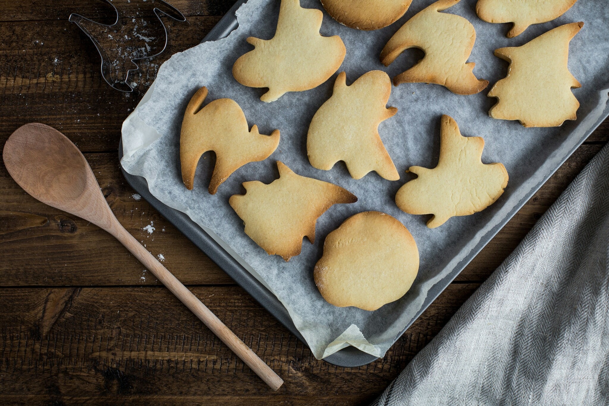 Halloween cat, ghost, and witch sugar cookies sit on a parchment paper lined baking sheet on a wooden countertop scattered with flour, a cookie cutter, a wooden spoon, and a white dish cloth. 