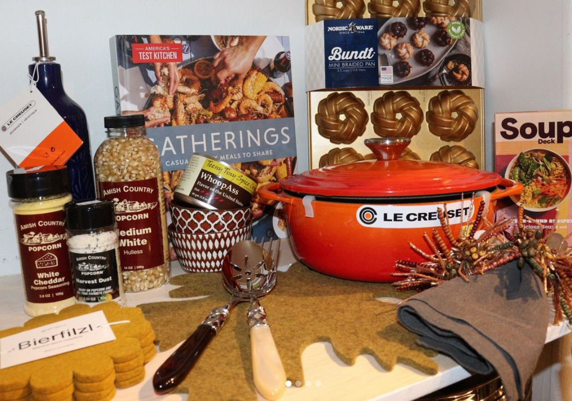 A collection of items, like an orange le creuset dutch oven, popcorn kernels, cook books, serving spoons, linen napkins, etc. arranged on a white tabletop