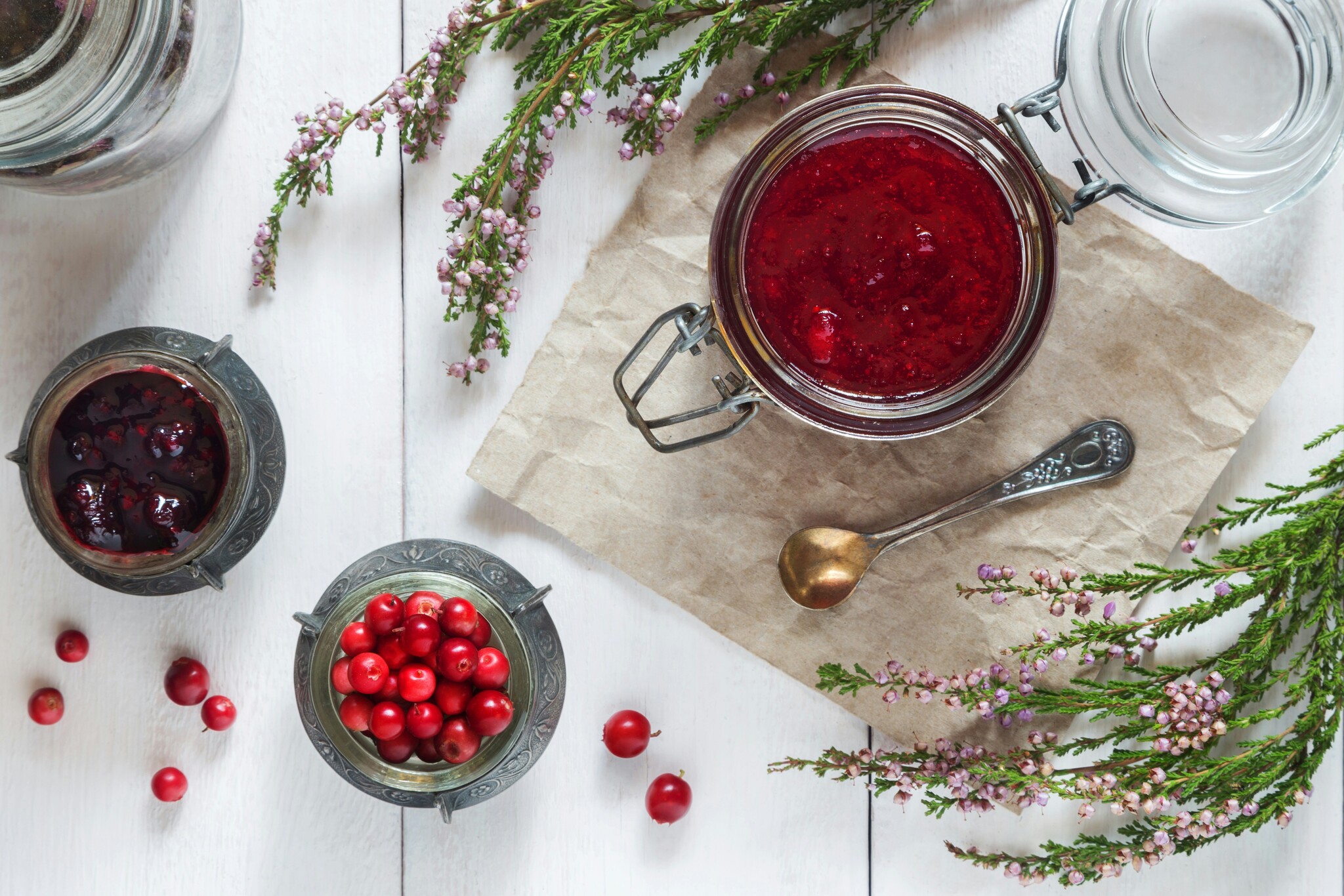 Cranberries and cranberry sauce sit in jars on a white tabletop with flowers and a spoon beside them. 