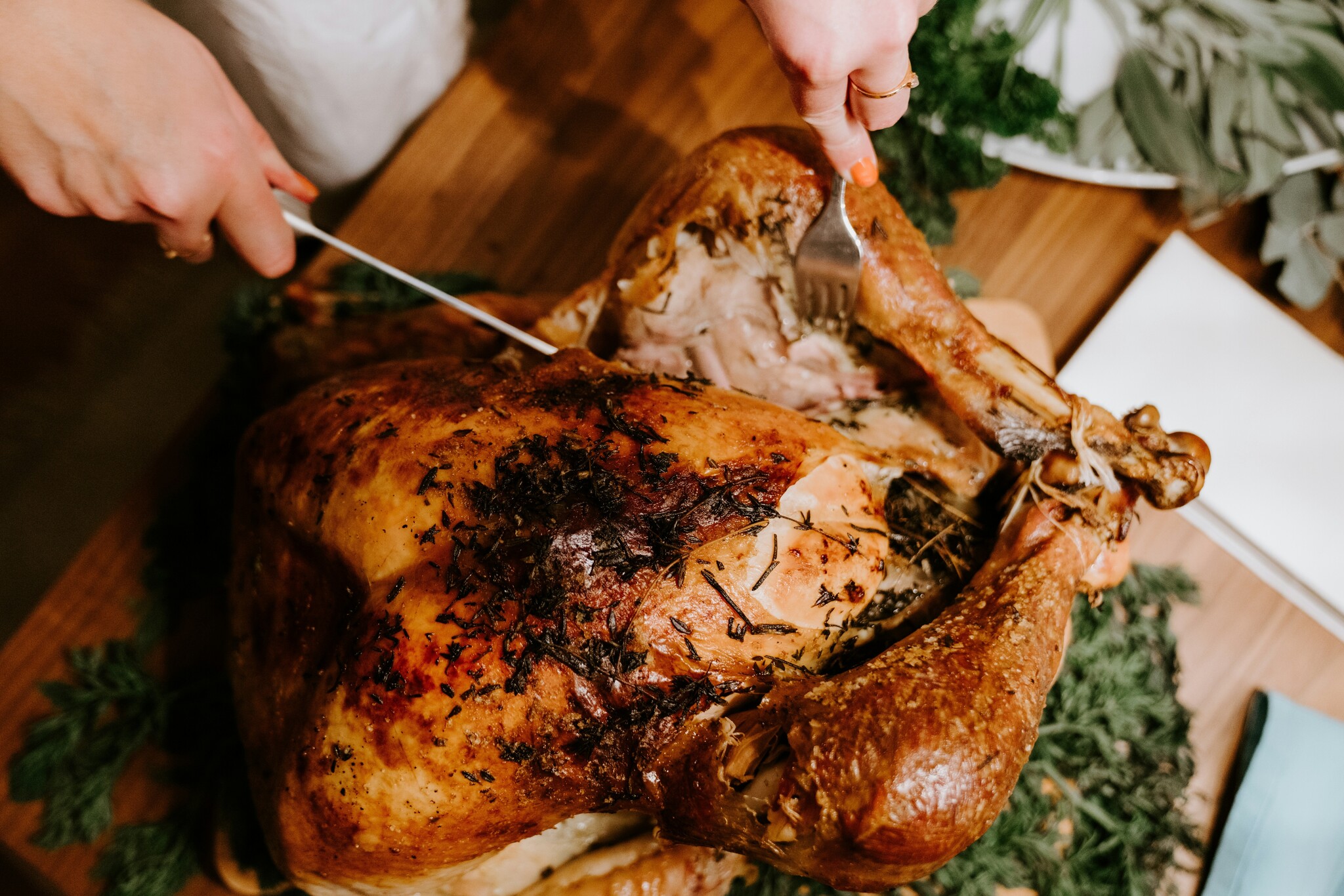 A woman cuts the leg off of a roasted Thanksgiving turkey on a wooden surface with herbs around it. 