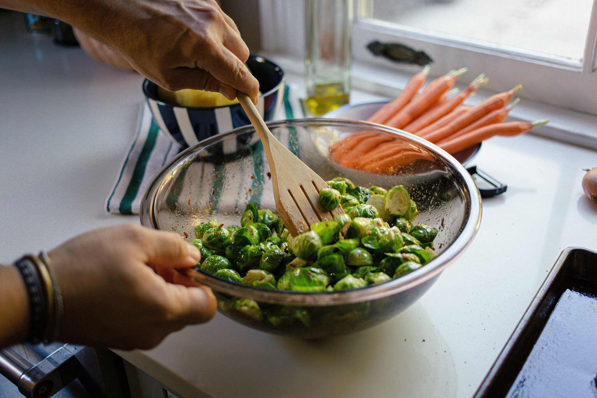 A man stirs brussels sprouts in a clear glass bowl on a white countertop with carrots in a bowl behind it. 