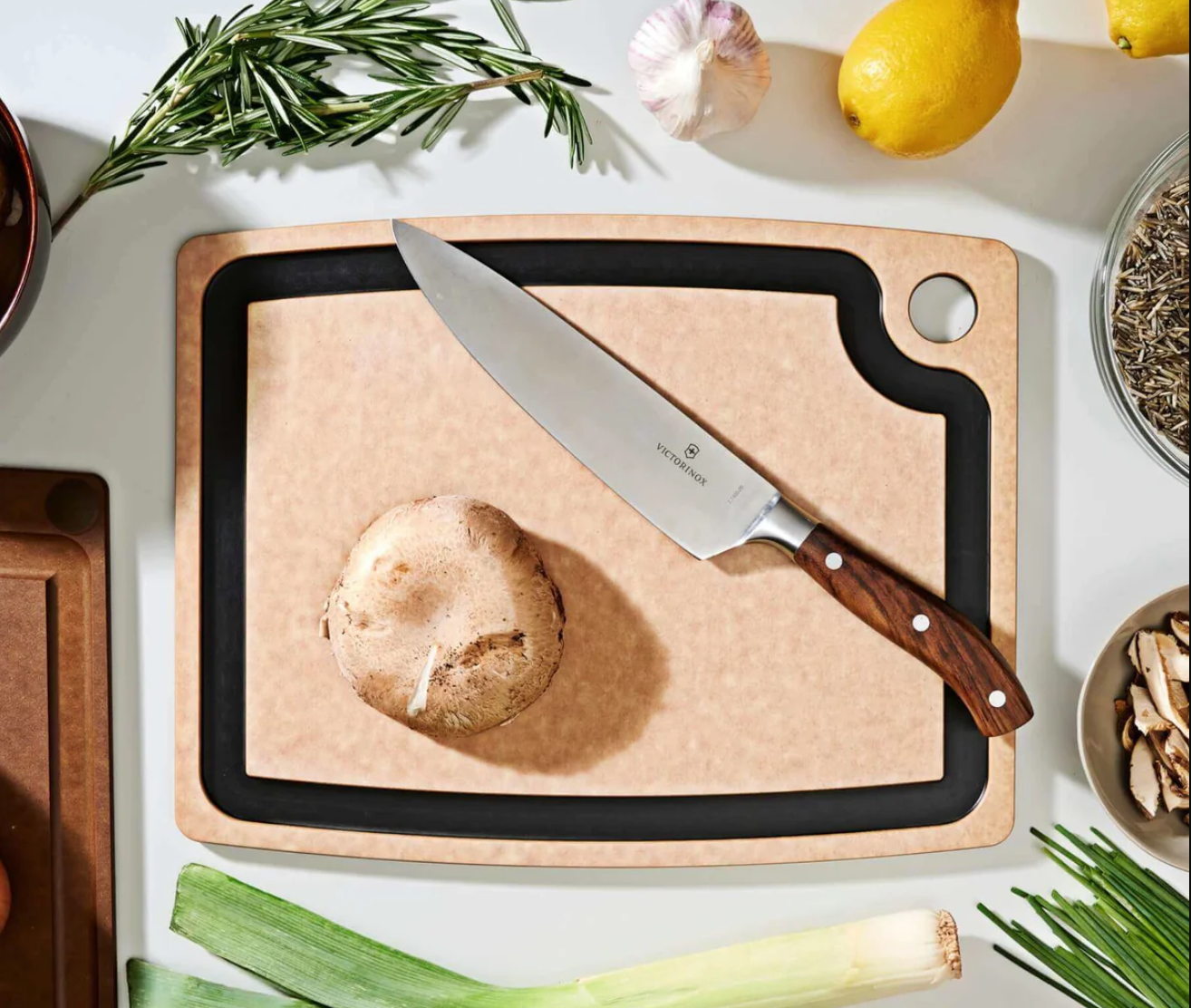 An epicurean cutting board with a juice groove sits on a white countertop surrounded by herbs, garlic, lemons, and leeks. On top of the cutting board rests a Victorinox knife and a mushroom. 
