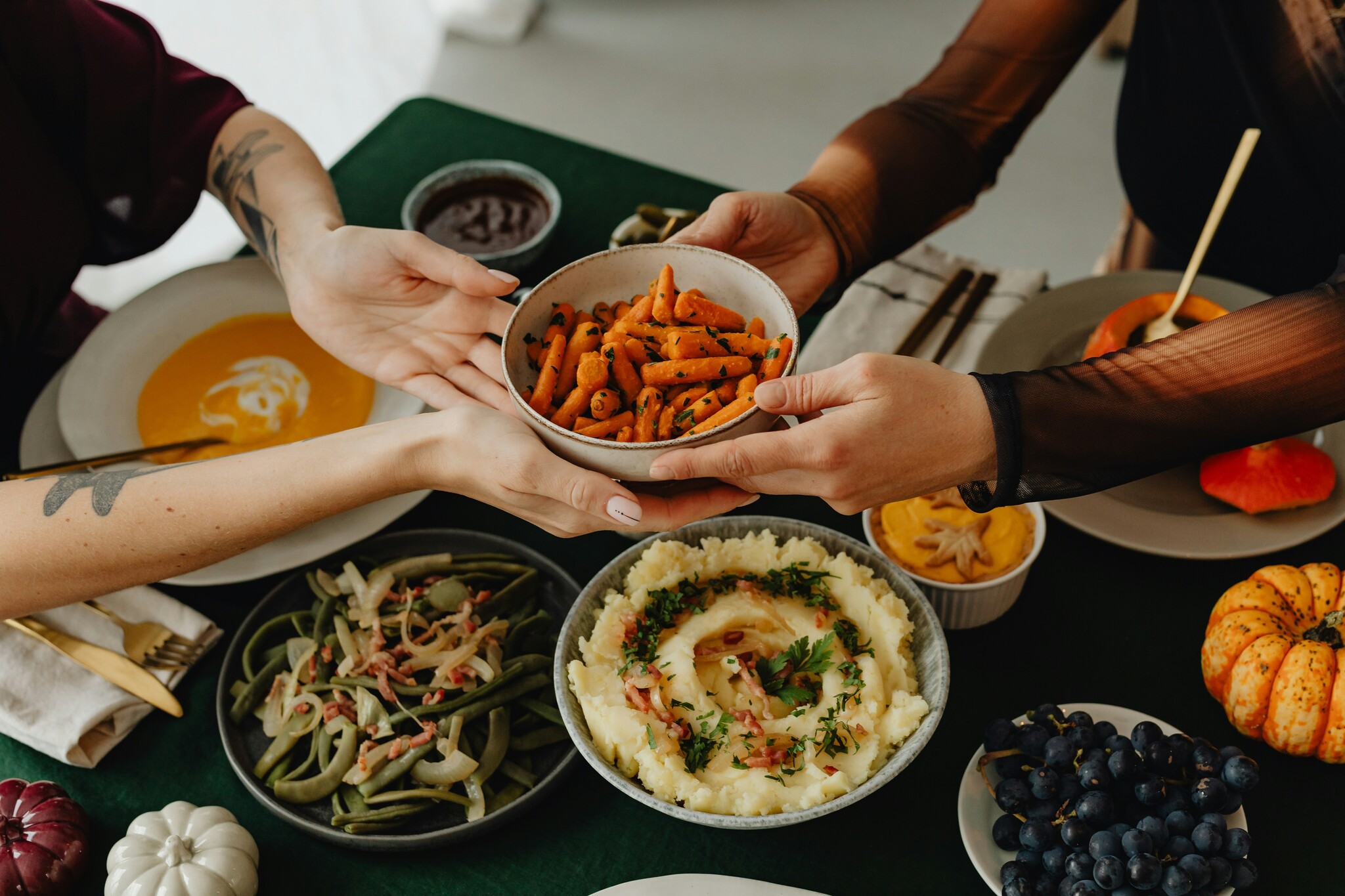 People pass a bowl of cooked carrots over a Thanksgiving spread consisting of butternut squash soup, mashed potatoes, fruits, and vegetables. 